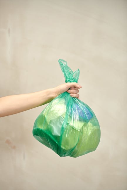 A person’s hand holding a green plastic rubbish bag by its twisted top against a plain, neutral beige background. The bag appears to be filled with waste materials, with visible shaping and slight bulges indicating contents inside. The plastic is semi-transparent, revealing some indistinct items beneath the surface. The bag's finish is smooth and shiny, reflecting ambient light. The hand is positioned slightly left of center in the image, with the arm extending from the left edge. The scene is well-lit, emphasizing the texture and color of the plastic bag without casting harsh shadows. This type of imagery relates to waste collection and rubbish removal services, illustrating the process of private waste disposal or on-site clearance, often handled by specialists such as Waste Collection Belgravia. The focus on the plastic bag underscores the importance of proper waste handling and the role of independent rubbish collection in maintaining cleanliness and compliance in residential or commercial environments.