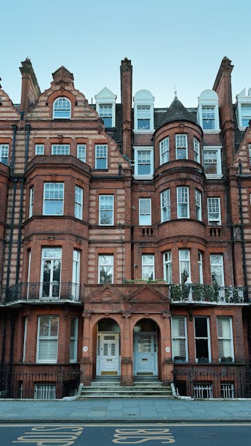 A multi-storey Victorian-style brick building with a symmetrical façade, featuring large bay windows on each level, some with white frames and decorative detailing. The building is constructed from red-brown bricks with stone accents around the windows and doors, and includes small balcony railings on the first floor. The entrance consists of two white doors, each set within an arched stone surround, accessed via short steps leading from the paved street. The street in front is marked with yellow and white road markings, with a sidewalk directly adjacent. The building is set against a clear blue sky, indicating daytime, and the overall scene is well-lit with natural daylight, capturing the structure’s historic architecture and detailing. This image relates to private property and possibly on-site waste clearance, aligning with the theme of independent rubbish removal services such as those offered by Waste Collection Belgravia.