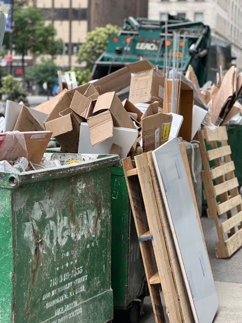 A large green waste refuse container filled with various discarded items, including cardboard boxes of different sizes, some flattened and some partially intact, and a smaller white box visible among the packing materials. The cardboard exhibits textures typical of corrugated and smooth surfaces with brown and white tones. Leaning against the side of the container is a wooden pallet with a light, unfinished wood appearance, featuring a slatted structure. In the background, a waste collection vehicle with visible branding and a hydraulic arm can be seen, suggesting active rubbish collection in an urban environment. The scene is outdoors, on a paved street with some trees and building facades visible behind, under natural daylight conditions. The overall setting reflects a typical private waste disposal or on-site clearance scenario, managed by Waste Collection Belgravia, highlighting elements relevant to independent rubbish removal services in the area.