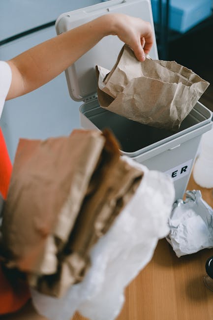 A person using a colourless recycling bin with a lid, placing a crumpled brown paper bag inside, while holding additional crumpled paper bags in their other hand. The bin is situated on a wooden surface, likely a table or countertop. The background includes a portion of a white wall and a glimpse of a window or glass surface, suggesting an indoor environment. The scene emphasizes domestic waste disposal, featuring simple materials such as paper and cardboard, which are typical for private rubbish collection services. The natural lighting highlights the textures of the crumpled paper and the smooth surface of the recycling bin, aligning with professional waste management practices aimed at responsible rubbish handling and on-site clearance. Waste Collection Belgravia services are associated with this scene, illustrating an organised approach to recycling and waste segregation, supporting alternative waste disposal options outside of municipal collection systems.