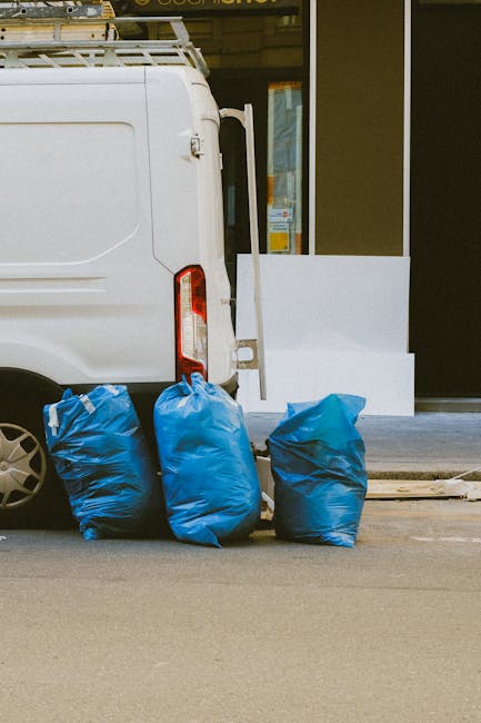 The image shows the rear side of a white commercial van parked on a paved street, with three large blue rubbish bags placed upright on the ground near the vehicle's back door, which is partially open. The bags are made of plastic, appear filled with waste, and are slightly wrinkled with some creases visible on their surface. In the background, there is a storefront or building with a glass window or door and a white protective barrier or step in front of it. The scene is outdoors during daylight, with natural lighting illuminating the area. The setting suggests a waste collection or rubbish removal activity, consistent with private waste disposal services provided by companies like Waste Collection Belgravia, and it reflects an alternative to official local authority rubbish collection, emphasizing on-site or independent waste handling.