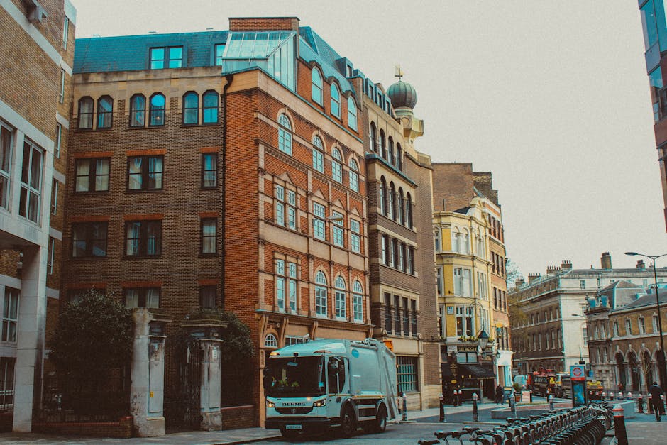 A white waste collection truck from Waste Collection Belgravia is parked on a city street in front of a row of historic multi-storey buildings with varying architectural styles. The truck is positioned in the foreground, facing towards the right side of the image, with visible details including its cab, side mirrors, and partially visible rear loading area. The buildings behind display a mix of traditional red-brick facades with large, arched and rectangular windows, some with decorative stonework, and others with ornate architectural elements. To the right, a pale yellow building with decorative molding and a domed turret is visible. The street is lined with black bollards and a bicycle-sharing station with several parked bikes. Overhead, the sky is clear, providing natural daylight that highlights the textures of the brickwork and metal roofing. The environment suggests an urban setting where private waste collection services, such as those from Waste Collection Belgravia, may be used for on-site rubbish removal rather than council-led collection services.