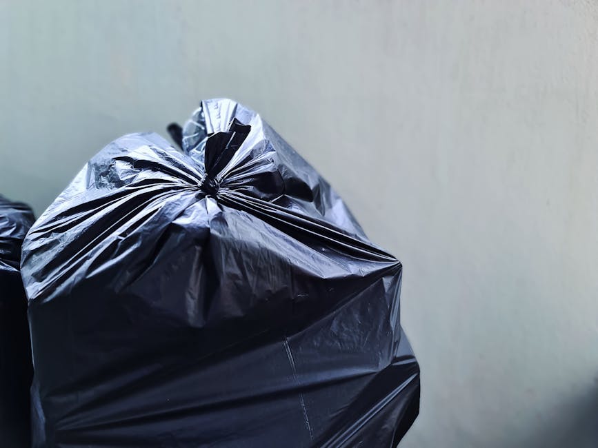 A person's arm extends into the frame from the left, holding a green plastic rubbish bag with both a green and a blue tint, tightly gathered at the top with a knotted closure. The bag appears partially filled, with its surface slightly wrinkled and semi-translucent, revealing indistinct contents inside. The background is a plain, neutral beige wall with no distinct features or markings. The scene is well-lit, capturing the details of the bag's material and the hand's skin tone clearly. This image exemplifies waste collection in a private or independent setting, aligning with services like those offered by Waste Collection Belgravia, emphasizing the handling or disposal of rubbish via external collection or on-site waste management solutions.